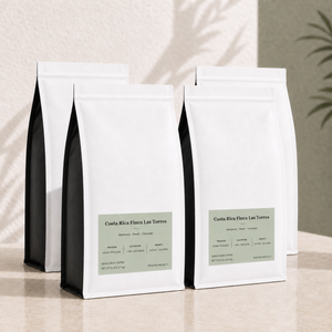 Four 5 lb white coffee bags with black side panels and pale green labels, arranged on a beige surface against a textured wall with soft shadow overlays. The front bags show the text Costa Rica Finos Las Torres.