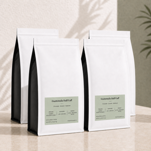 Four bulk 5 lb white coffee bags with black sides and pale green labels,  "Guatemala Half Caff" standing on a light beige surface against a textured beige wall with soft shadow overlays.