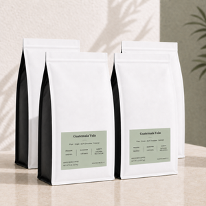 Four white bulk 5 lb coffee bags with black sides and a muted sage green label reading 'Guatemala Yula' on a light beige surface against a textured beige wall with soft leaf shadows.