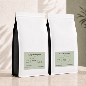 Two white bulk 5 lb coffee bags with black side panels and pale sage green labels, arranged on a light beige surface against a softly textured beige wall with leaf shadows. Kenya Kiamugumo