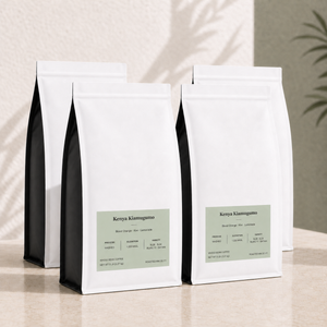 Four white bulk 5 lb coffee bags with black side panels and pale sage green labels, arranged on a light beige surface against a softly textured beige wall with leaf shadows. Kenya Kiamugumo